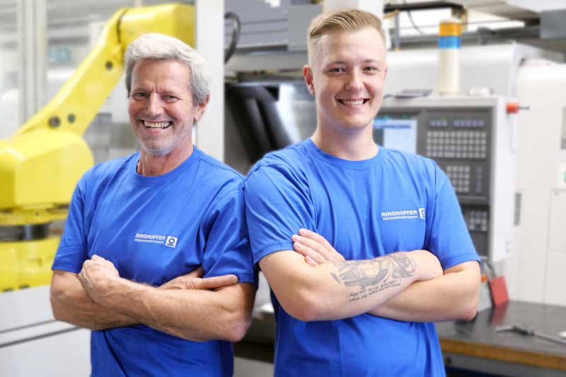 Zwei männliche Mitarbeiter stehen Schulter an Schulter mit verschränkten Armen in der Werkstatt und lächeln freundlich in die Kamera
